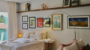 A well-lit bedroom with a bed, bedside table, and a lamp. There are framed artworks on the wall and a window with curtains. - Loft Rua Direita Você vai Adorar Este Lugar Único e Romântico (Centro Pirenópolis)