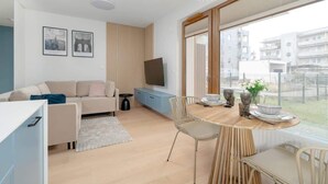 Ein modernes Wohnzimmer mit einer Couch, einem Esstisch mit Stühlen, einem an der Wand befestigten Fernseher und einem großen Fenster mit Blick auf die Stadt.