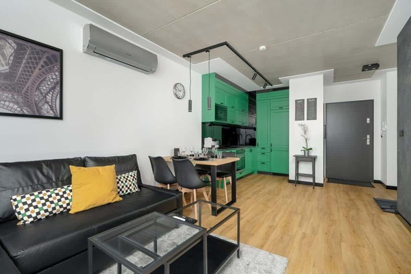 A modern living area with a black sofa, warm wooden flooring, and bold green cabinets in the background. The open layout seamlessly combines the lounge and kitchen spaces.