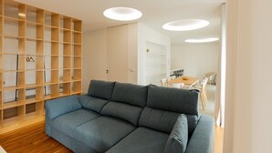 A modern living room with a grey sofa, wooden shelving unit, and a dining area with a table and chairs.