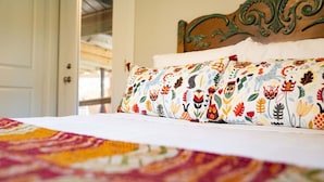Ein Bett mit einem farbenfrohen Quilz und einem gemusterten Kopfkissen, ein hölzernes Kopfende und eine weiße Tür im Hintergrund.