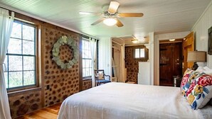 A bedroom with a large bed, a ceiling fan, a stone-patterned wall, and a window with curtains. - Rustic Retreat: Tub, Firepit, Porch Swings & Pool (San Marcos)