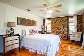 Quarto com piso de madeira, cama com roupa de cama branca, mesa de cabeceira, luminária, cômoda, ventilador de teto e parede com padrão de pedra.