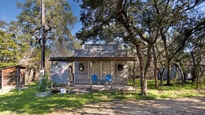 A rustic cabin with a porch, a small shed, and a large tree in the background. - Outpost Vibes: Outhouse, Outdoor Shower & Pool (San Marcos)