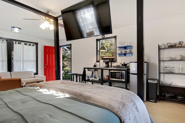 A bedroom with a bed, a TV mounted on the wall, a desk with a chair, and a window with a view of greenery.