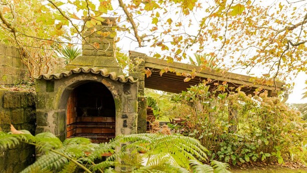 Un rústico edificio de piedra con chimenea de ladrillo, rodeado de exuberante follaje y helechos.