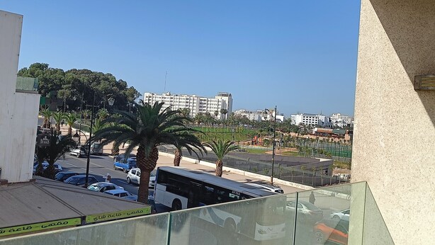 Vista desde un balcón sobre un estacionamiento con varios coches, un autobús y palmeras. Al fondo, edificios de varios pisos y un cielo azul despejado.