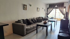 Una sala de estar con un sofá gris, una mesa de centro y un televisor. Hay dos cuadros enmarcados en la pared y una ventana con cortinas.