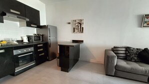 Una cocina moderna con armarios oscuros, microondas integrado y refrigerador de acero inoxidable. En la zona de estar, hay un sofá gris con cojines estampados.