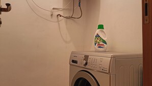 Una lavadora de carga frontal con panel de control Samsung, un calentador de agua montado en la pared y una botella de detergente Ariel sobre la máquina.