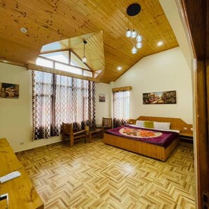 A room with a wooden ceiling, a bed with a floral bedspread, a dining table with chairs, and patterned curtains. - Hotel Jamla Regency Manali (Manali)