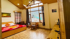 A bedroom with a wooden ceiling, a bed with a colorful blanket, a desk with a computer, and a window with a view of a house and mountains. - Hotel Jamla Regency Manali (Manali)