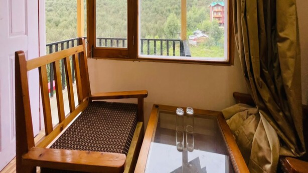 A room with a wooden chair, a small table with glasses, and a large window offering a view of a mountainous area. - Hotel Jamla Regency Manali (Manali)