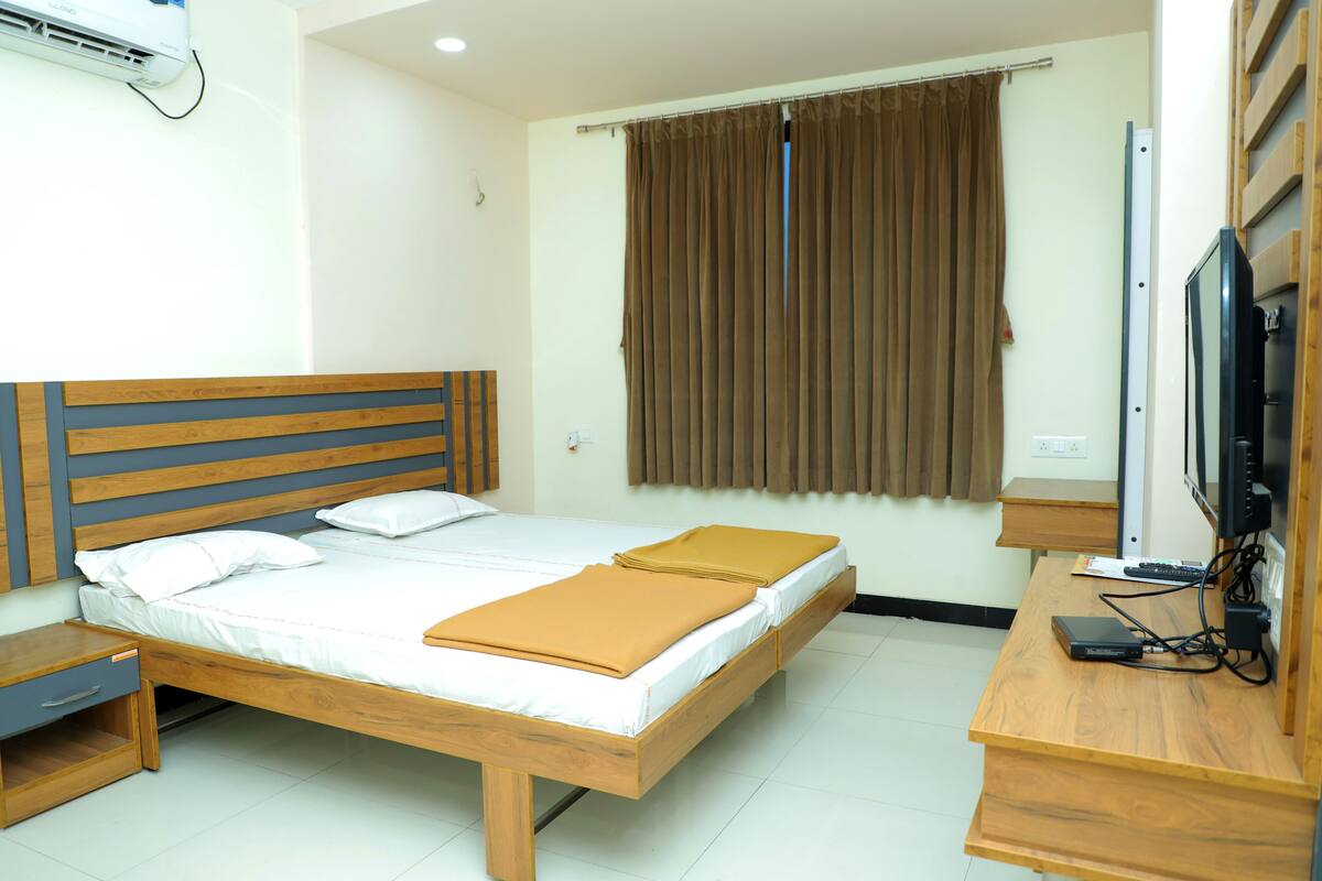A double bed with a wooden headboard, a bedside table, a desk with a computer and a TV, and a window with curtains.