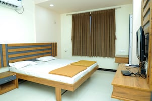 Uma cama de casal com cabeceira de madeira, uma mesa de cabeceira, uma escrivaninha com um computador e uma televisão, e uma janela com cortinas.