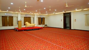 A spacious hall with a red carpet, multiple ceiling fans, and a stage with a podium and a sofa. - Hotel Anjani  (Latur)