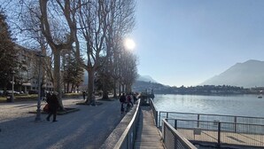 Une promenade en bois longe une rivière, où l’on peut voir des gens se promener et s’asseoir sur des bancs. Des arbres bordent le sentier, et une montagne est visible au loin.