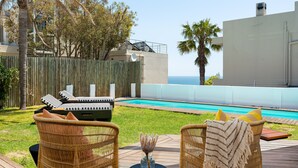 A deck with wicker chairs, a small table, and a pool in the background. - Neighbourgood First Crescent (Cape Town)