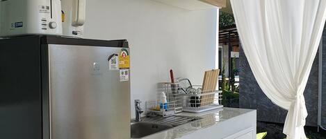 A modern kitchen with a stainless steel refrigerator, a built-in oven, and a microwave. There is a sink with a dish rack and a window with white curtains.
