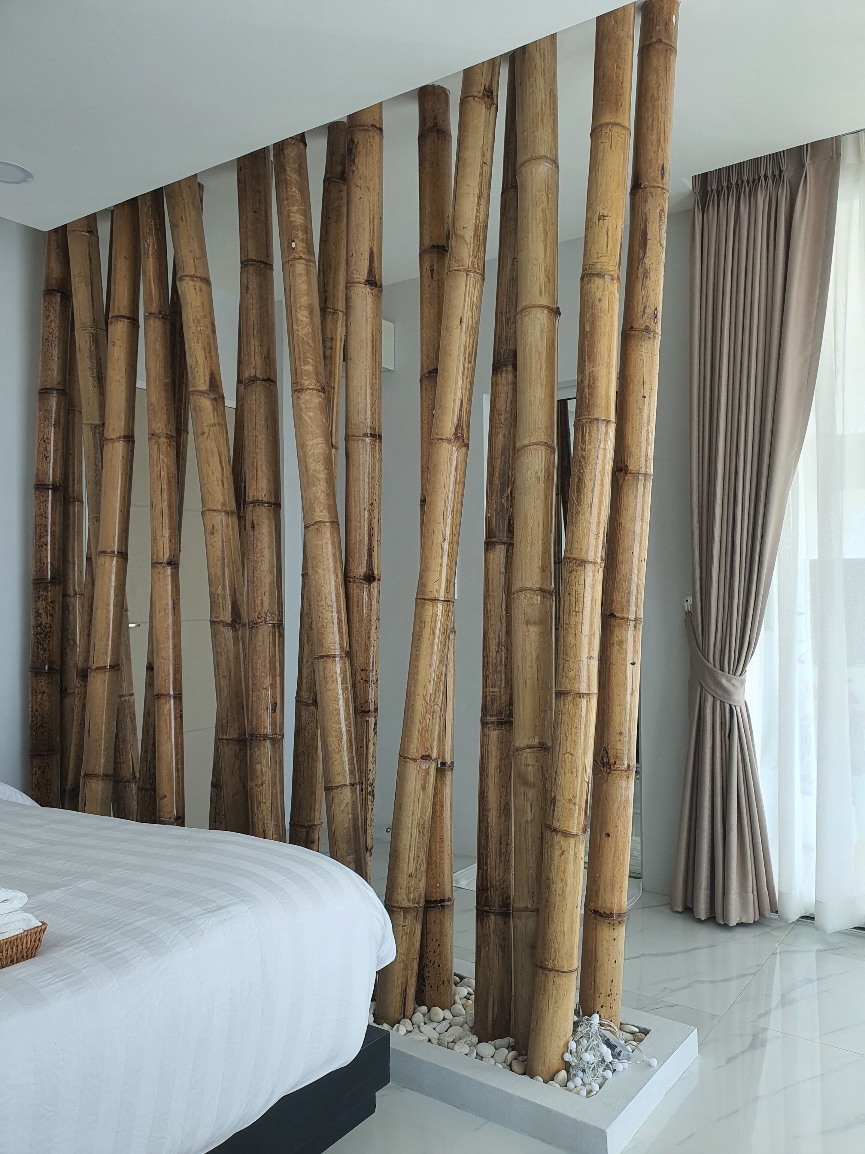 Un dormitorio moderno con una cama grande, un separador de ambientes de bambú y vistas al exterior a través de una ventana con cortinas.