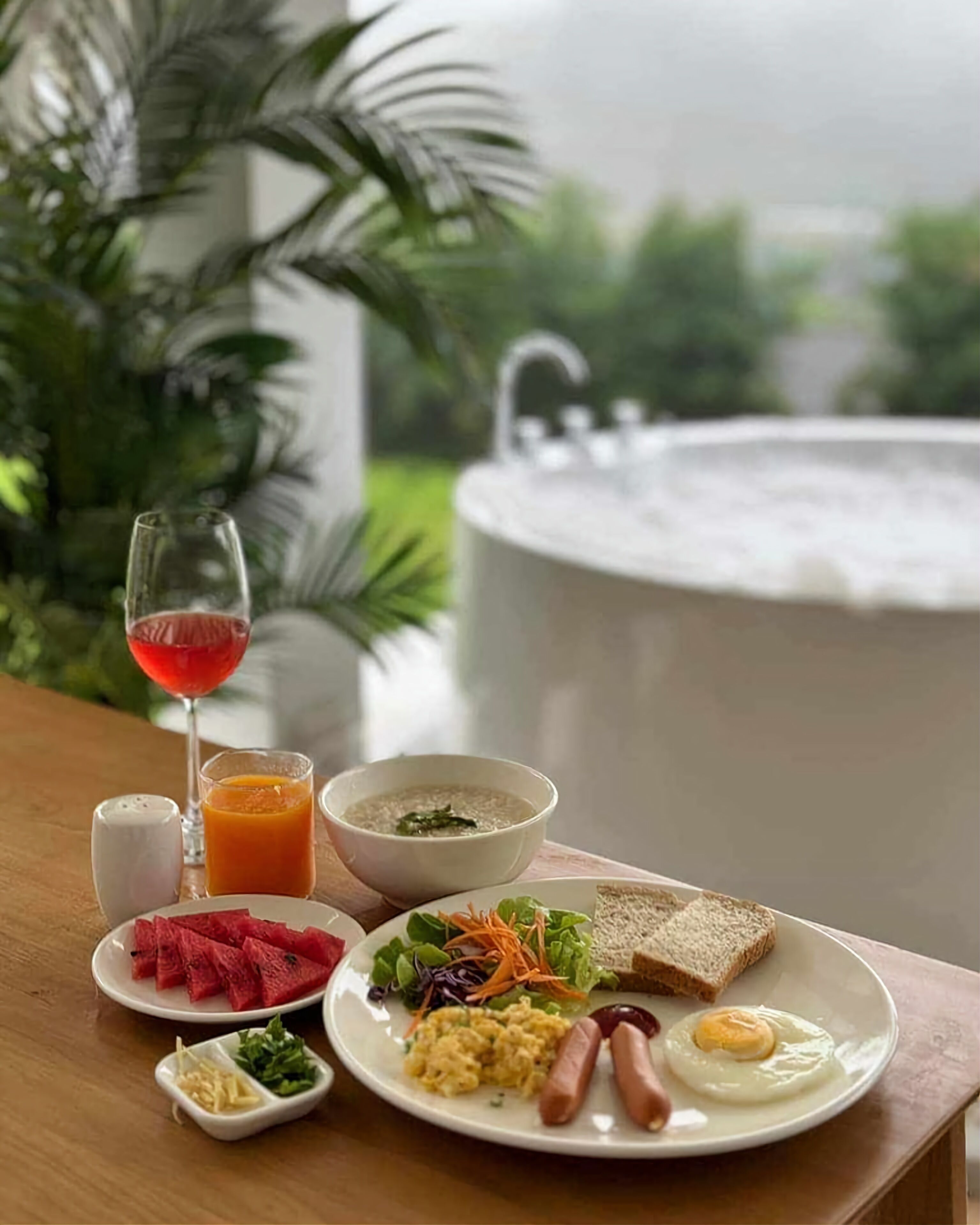 A breakfast table with a variety of food items including toast, eggs, sausages, and a salad, along with a glass of red juice and a bowl of soup.