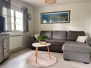 Olohuoneessa on harmaa sohva, pieni pyöreä pöytä, jossa on kasvi, televisio, joka on sijoitettu kaappiin, ja seinällä on kehystetty taulu.