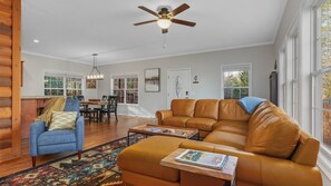 Uma sala de estar ampla com um sofá de couro marrom, uma poltrona azul e uma mesa de centro. Há um ventilador de teto, uma área de jantar com mesa e cadeiras de madeira e grandes janelas que proporcionam iluminação natural.