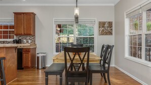 Une cuisine avec des meubles de rangement en bois, une table à manger avec des chaises, une poubelle et une fenêtre donnant sur un espace verdoyant.