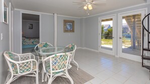 A spacious room with a glass table and white wicker chairs, a ceiling fan, and a view through a glass door to an outdoor area.