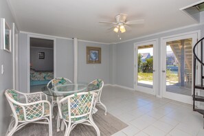 A spacious room with a glass table and white wicker chairs, a ceiling fan, and a view through a glass door to an outdoor area.