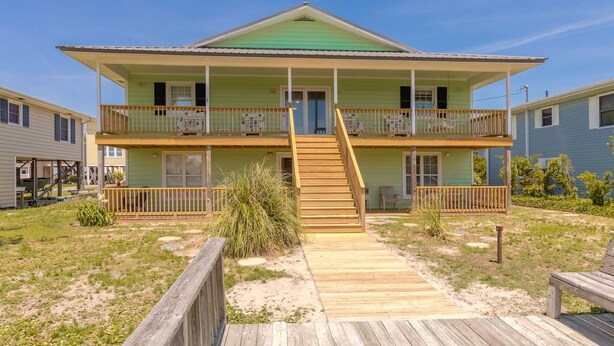 A two-story beachfront house with a wooden deck leading to the front door, a balcony on each floor, and a clear view of the ocean.