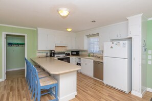 A compact kitchen with white cabinets, a central island with blue chairs, a refrigerator, and a microwave. - A Leave of Absence - 5BR Canalfront Home with Covered Porches and Boat Dock (Topsail Beach)