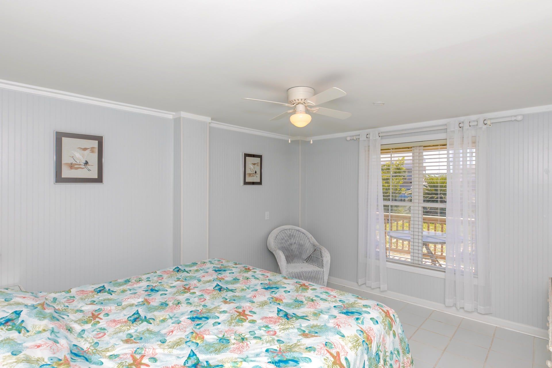 A bedroom with a bed, a ceiling fan, two framed pictures, a wicker chair, and a window with blinds.