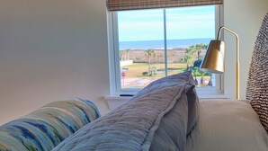 A bedroom with a view of the ocean, a bed with a striped pillow, a lamp, and a woven headboard. - Ultimate Beach Escape -4BR Luxury with EV Charging (Galveston)