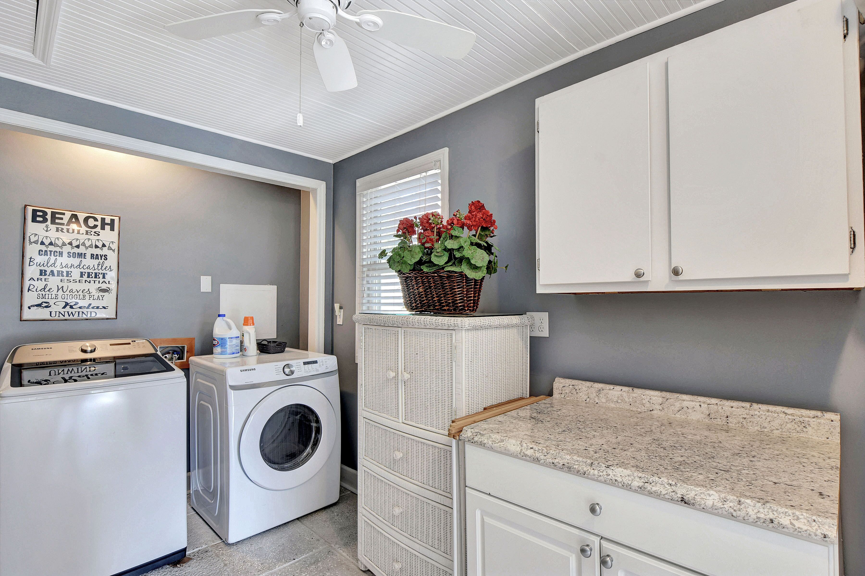 A laundry room with a washing machine, dryer, and a ceiling fan. There is a wall-mounted basket with flowers and a sign with text.