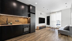 A modern kitchen with dark cabinetry, a wooden herringbone backsplash, and a built-in oven. There is a dining area with a grey sofa and a small table. - RentPlanet - Apartamenty Wiktoryn (Warsaw)