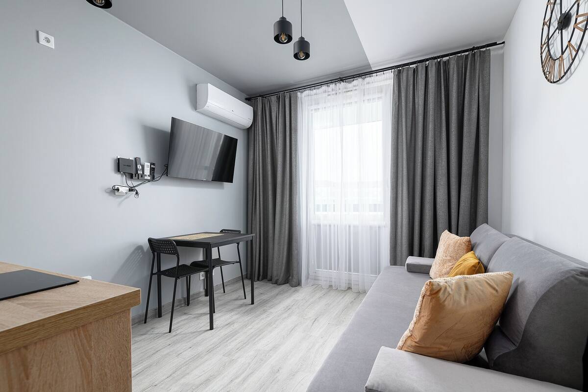 A modern living room with a grey sofa, a wooden desk, and a TV mounted on the wall.
