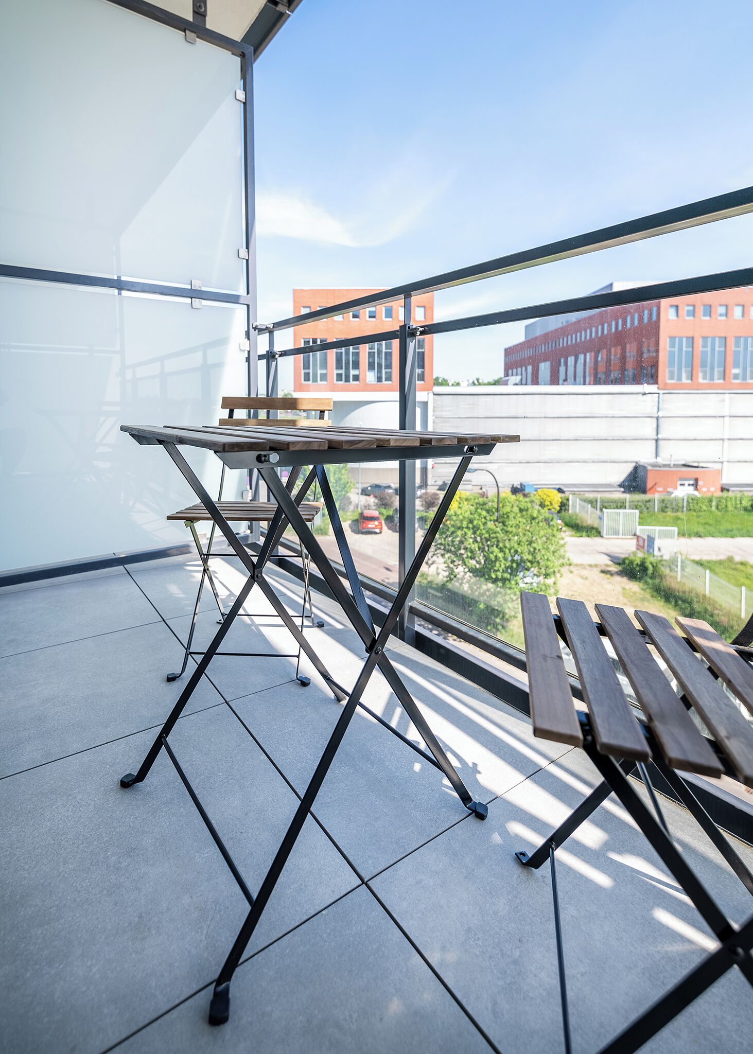 An outdoor balcony with a wooden table and benches, a metal railing, and a view of buildings in the background.