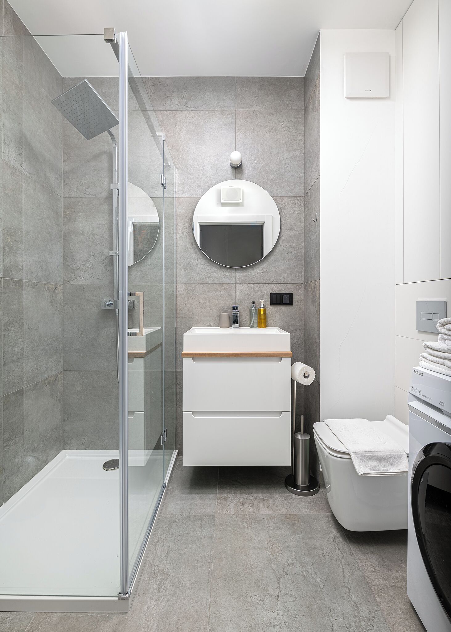 Une salle de bain moderne dotée d’une douche à parois de verre, d’un miroir rond, d’un lavabo blanc avec un plan de toilette en bois et de toilettes intégrées.