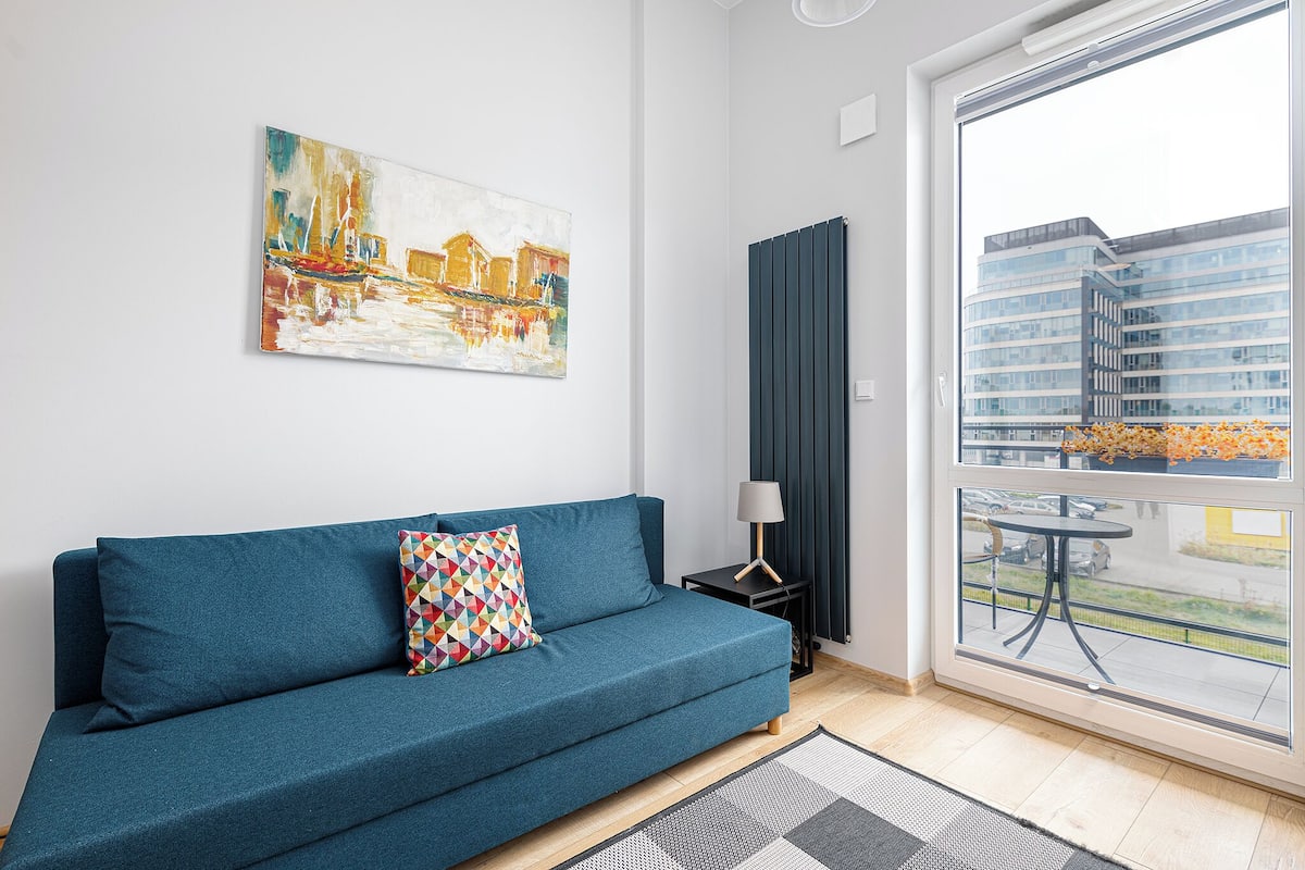 A modern living room with a blue sofa, a colorful geometric pillow, a painting of a cityscape, a small side table with a lamp, and a view of a building through a large window.