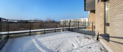 A rooftop terrace with a snow-covered ground, a glass balustrade, and a view of a distant building.