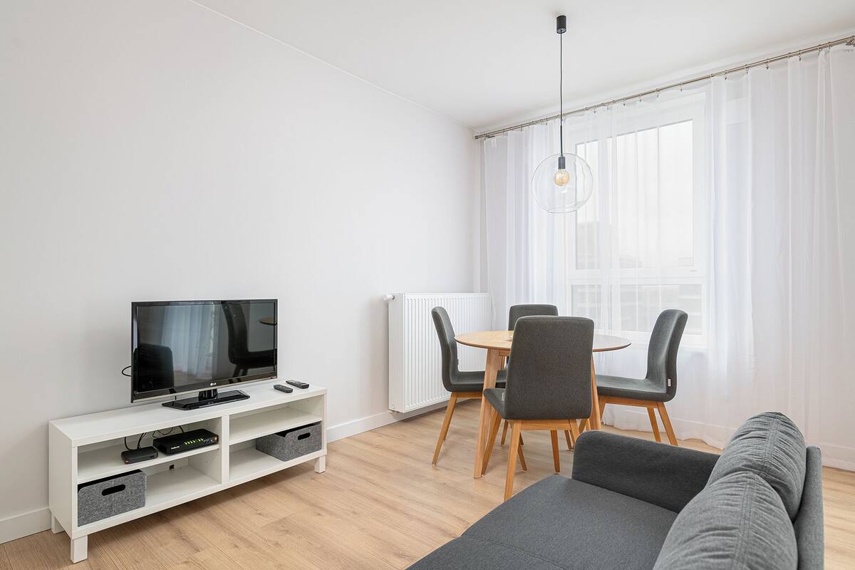 A modern living room with a flat-screen TV, a white entertainment unit, a grey sofa, a dining table with chairs, and a large window with sheer curtains.