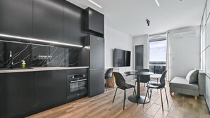 A modern kitchen with black cabinetry, a marble backsplash, and a built-in oven. There is a dining area with a round table and chairs, and a grey sofa with a view of the outdoors. - RentPlanet - Apartamenty Wiktoryn (Warsaw)