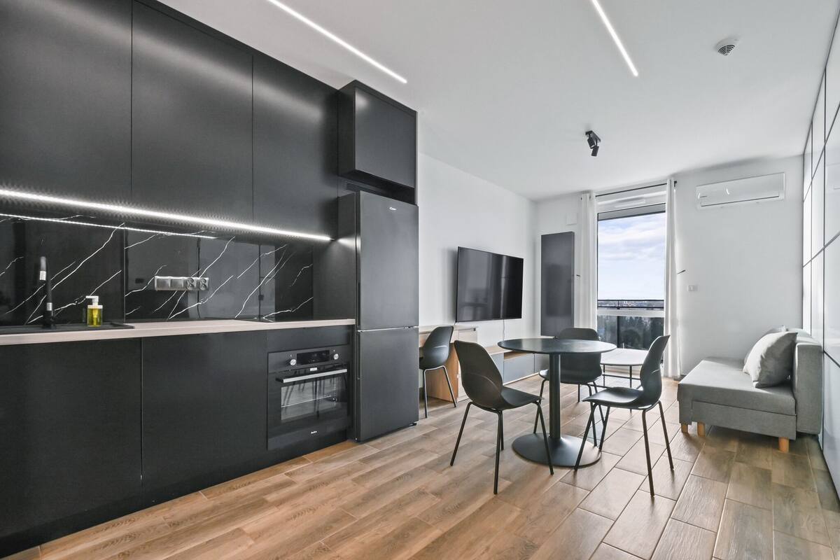 A modern kitchen with black cabinetry, a marble backsplash, and a built-in oven. There is a dining area with a round table and chairs, and a grey sofa with a view of the outdoors.