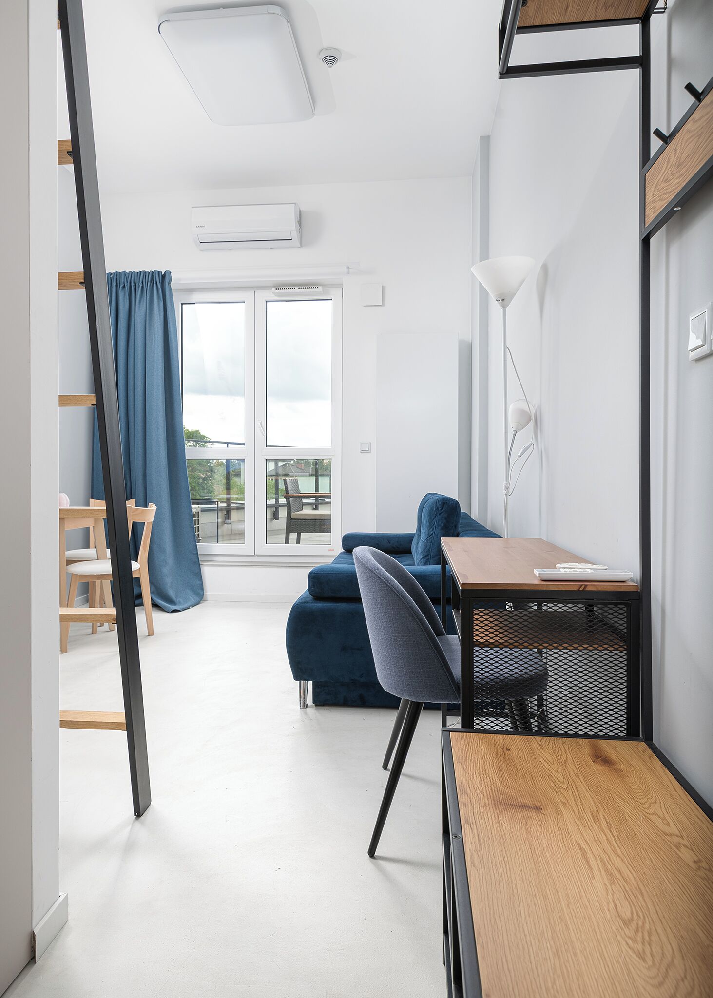 A modern, compact room with a wooden desk, a blue sofa, and a dining area with chairs.
