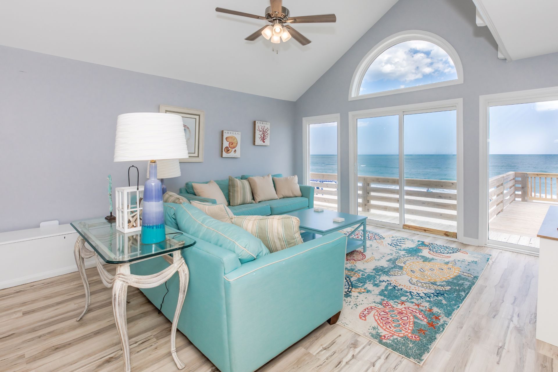 A spacious living room with a large window overlooking the ocean, a ceiling fan, and a light-colored wooden floor.