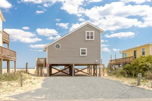 Une maison de plage à deux étages, dotée d’un balcon, d’une terrasse en bois et d’une allée gravillonnée.