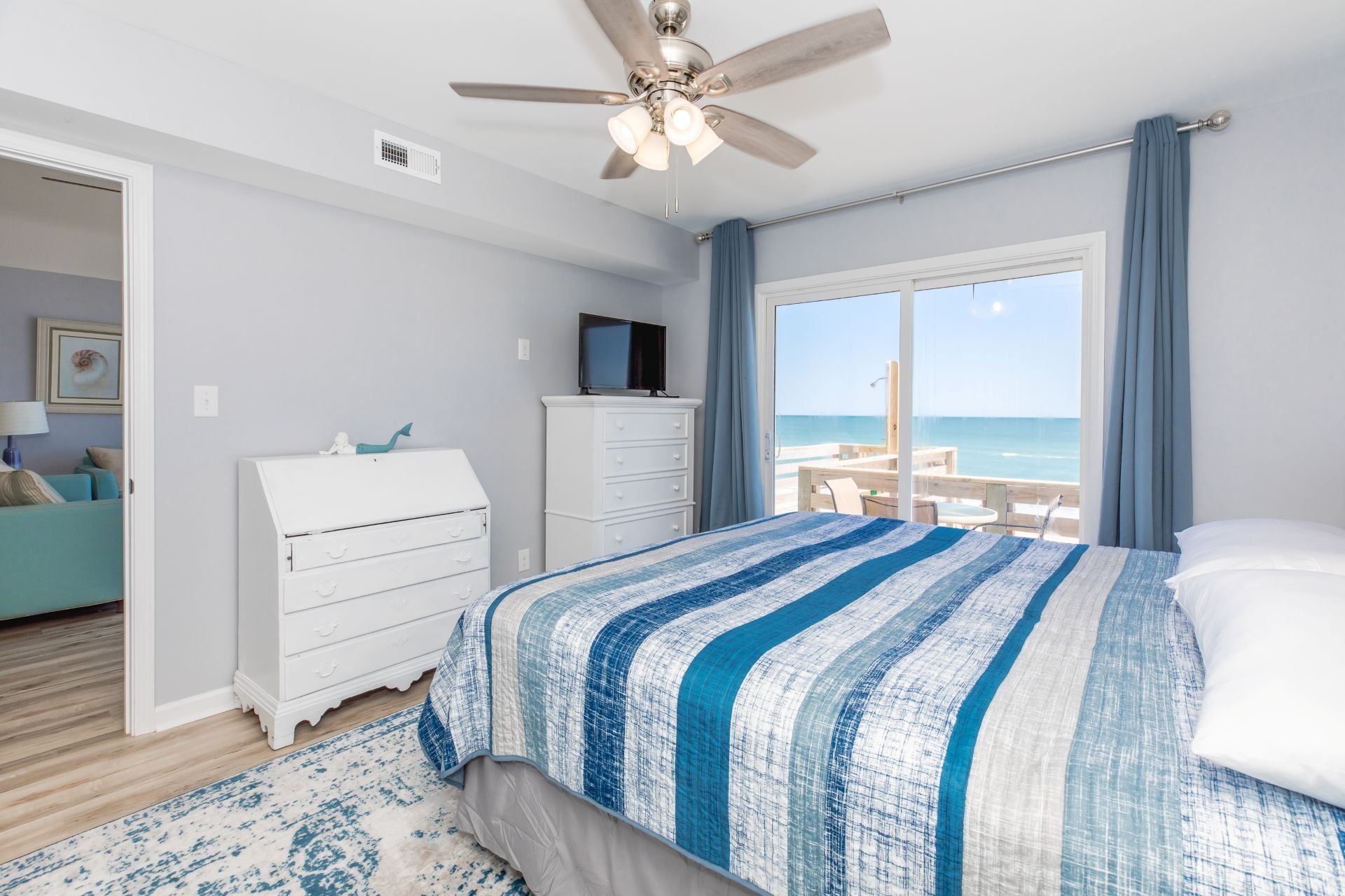 A bedroom with a bed, a ceiling fan, a dresser, a television, and a view of the ocean.