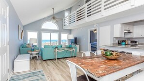 A spacious living area with a high ceiling, a dining table with a rustic wooden top, a teal sofa, and a view of the beach through large windows. - Sea Glass - Fantastic Views from this 3 BR Oceanfront Home! (Surf City)
