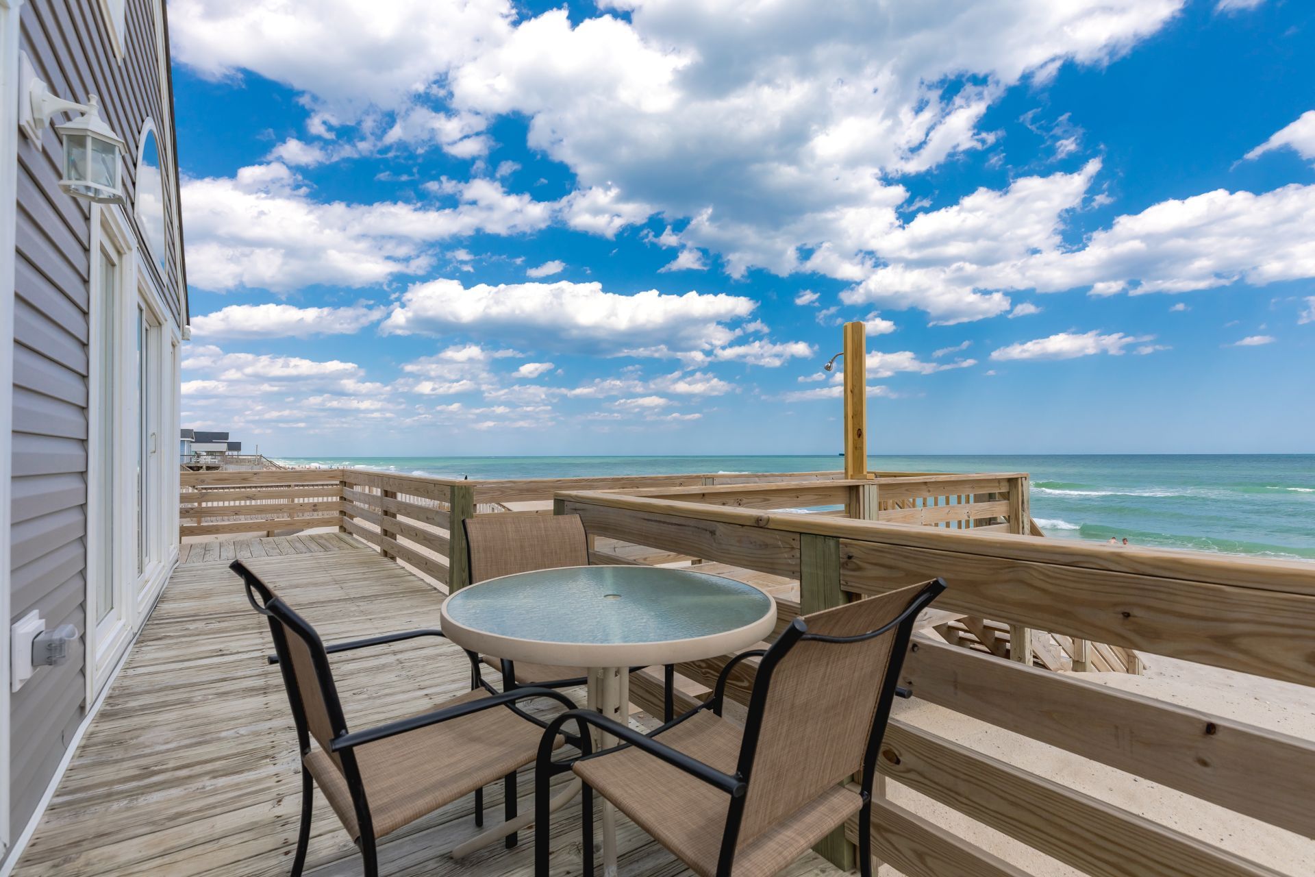 Una terraza de madera con una mesa redonda y sillas, con vista a una playa que cuenta con barandilla de madera y cielo despejado.